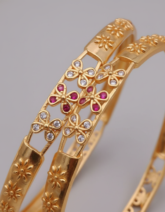 Gold bangles with floral designs and red gemstones on a gray background
