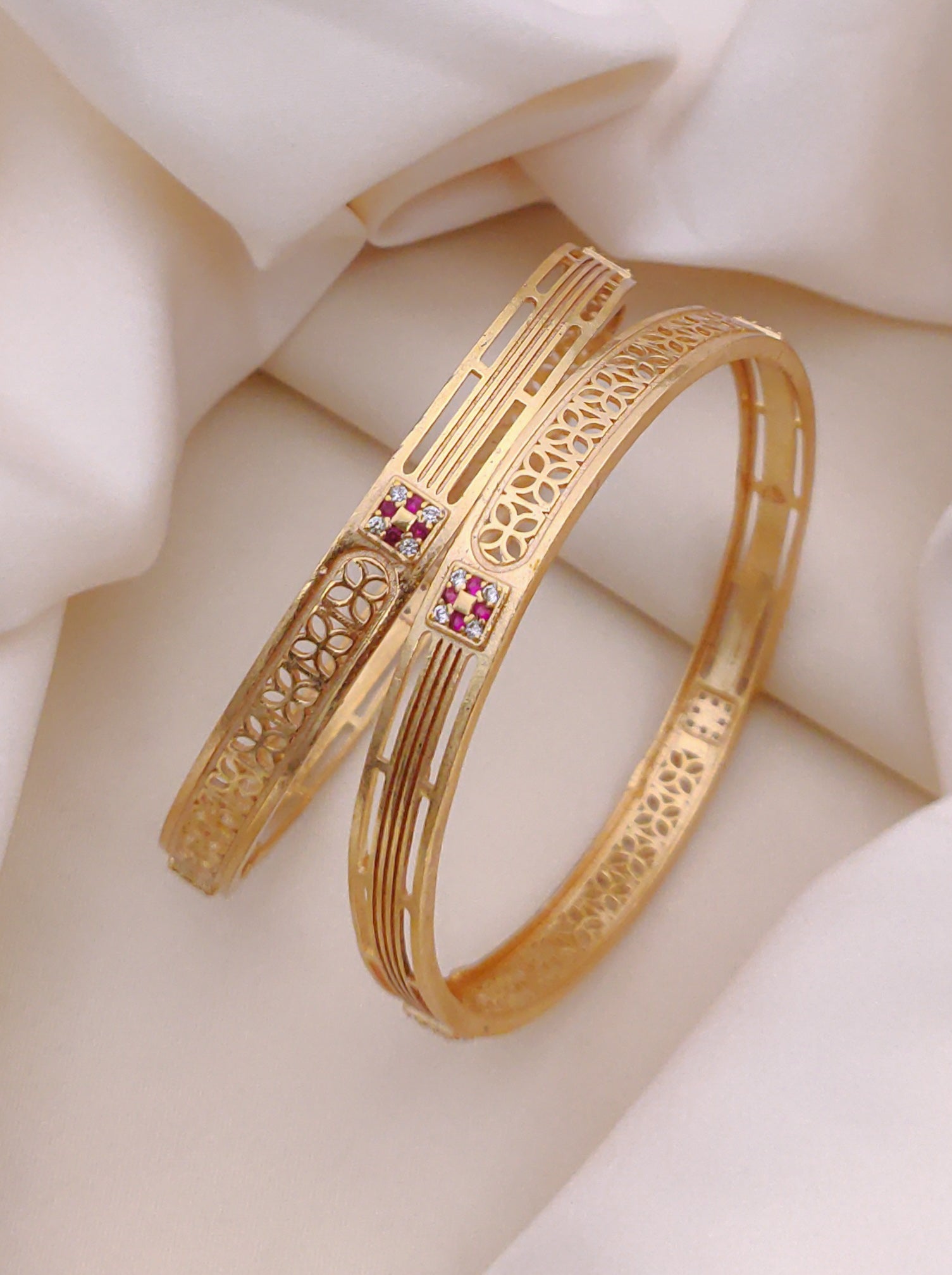 Gold bangles with intricate designs and small pink stones on a soft beige fabric background