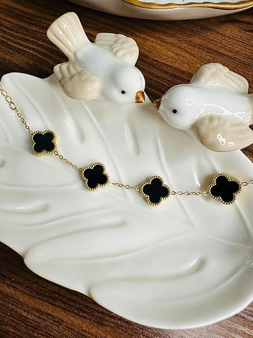 Gold necklace with black clover-shaped pendants on a white ceramic dish with birds.