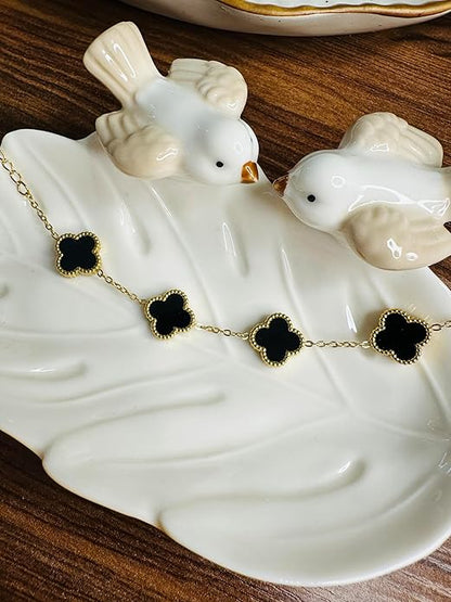 Gold necklace with black clover-shaped pendants on a white ceramic dish with birds.